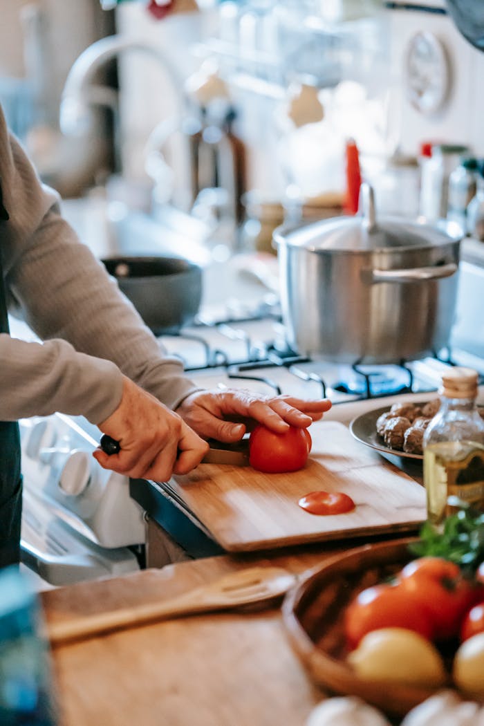 Crop unrecognizable person cutting ripe tomato while preparing lunch against gas stove in house