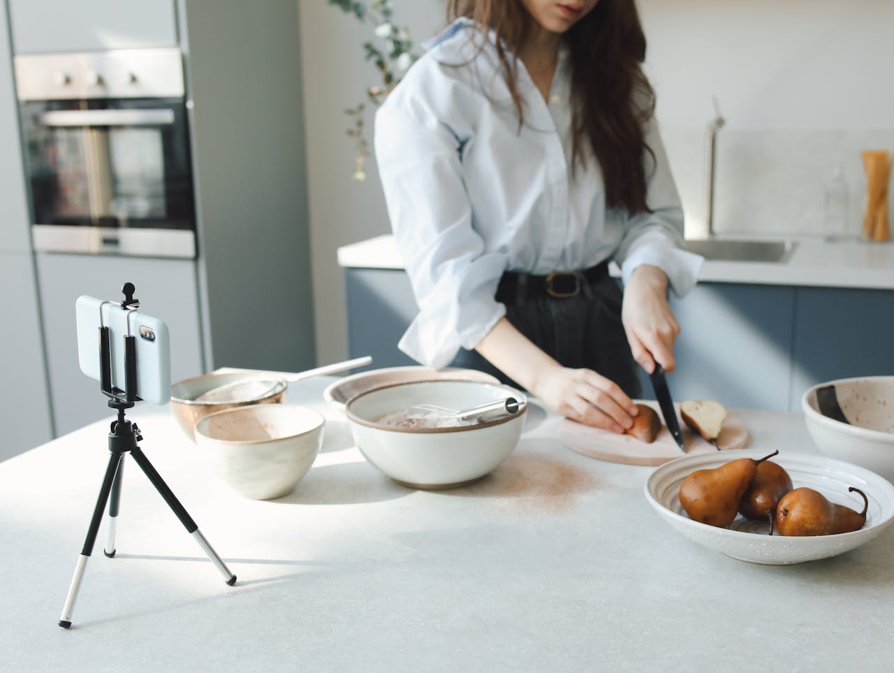 Woman slicing bosc pears in a modern kitchen, capturing the process on smartphone.