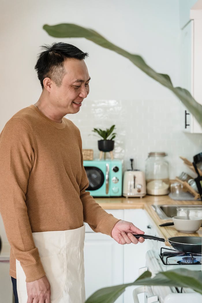 Smiling Asian man cooking in a stylish kitchen with modern appliances and natural light.