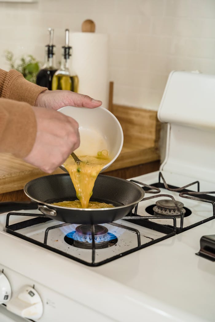 Close-up of hands pouring scrambled eggs into a pan on a gas stove, cooking breakfast.